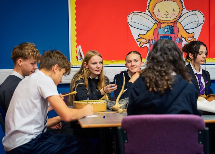 A group of secondary school children sit around a table. They are wearing blue jumpers and white shirts. They are actively engaged in conversation and making notes. They are immersed in the task, appear focused yet relaxed.